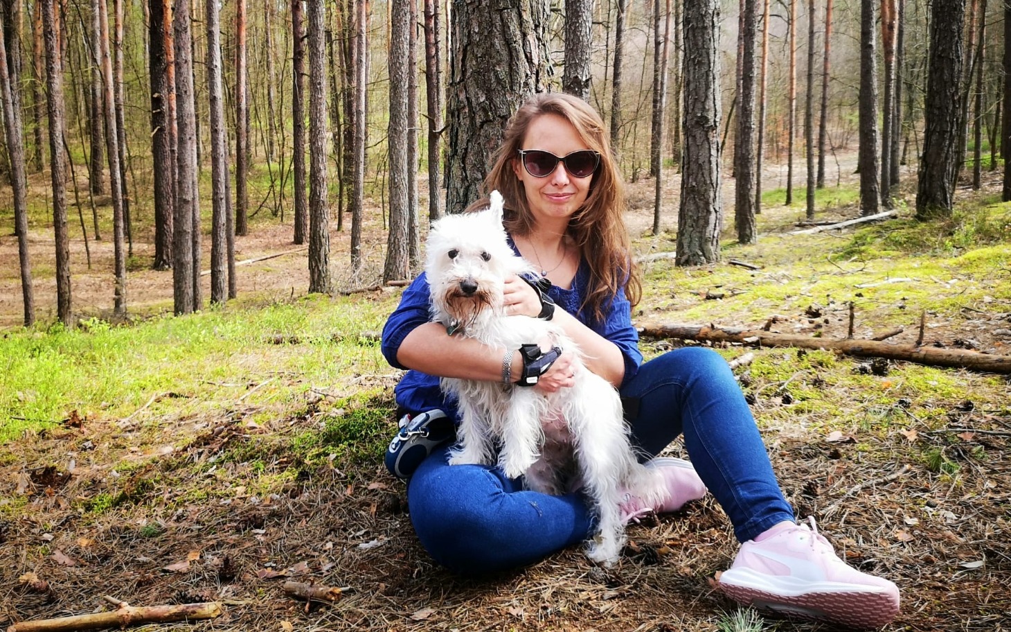 
        Kobieta siedząca na trawie z psem w lesie.
    