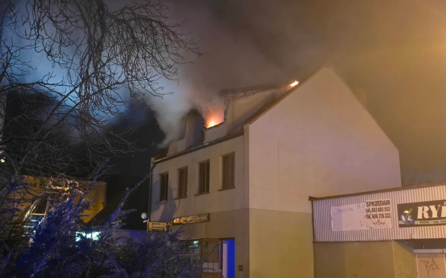 
        Płonący dach budynku w nocy, z dymem unoszącym się w powietrzu.
    