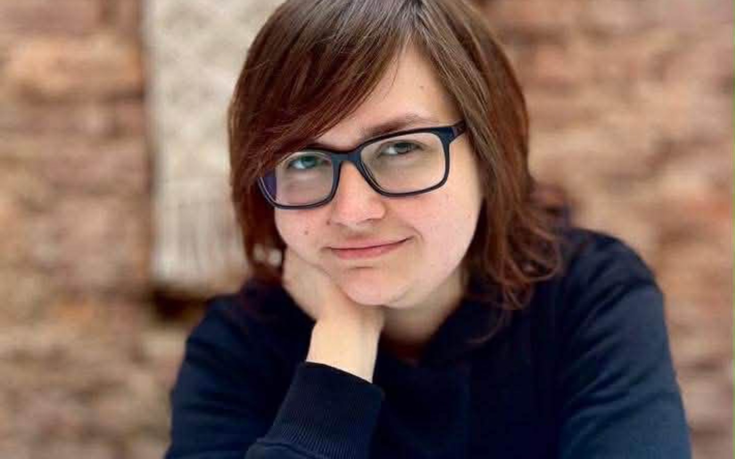 
        Portret młodej kobiety z uśmiechem na tle ceglanej ściany.
    