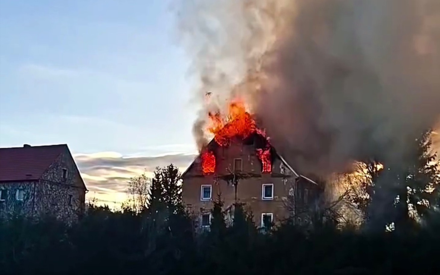 
        Zniszczony dom w płomieniach, dym unoszący się w powietrzu.
    