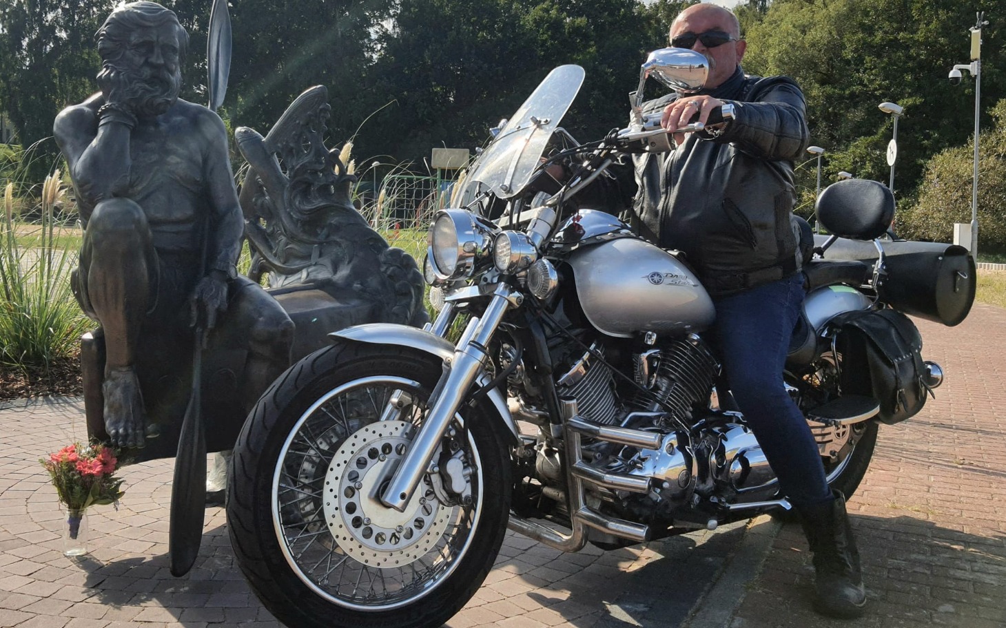 
        Mężczyzna na motocyklu, pozujący obok rzeźby w parku.
    