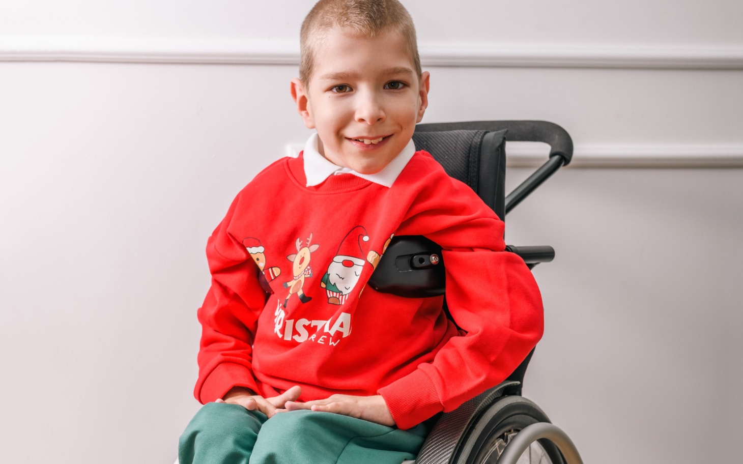 
        Chłopiec na wózku inwalidzkim, uśmiechający się w czerwonym swetrze.
    