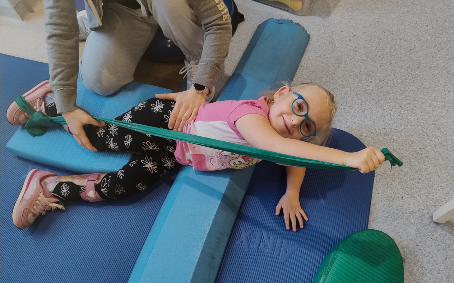 
        Dziewczynka ćwiczy z pomocą terapeuty, uśmiechnięta.
    