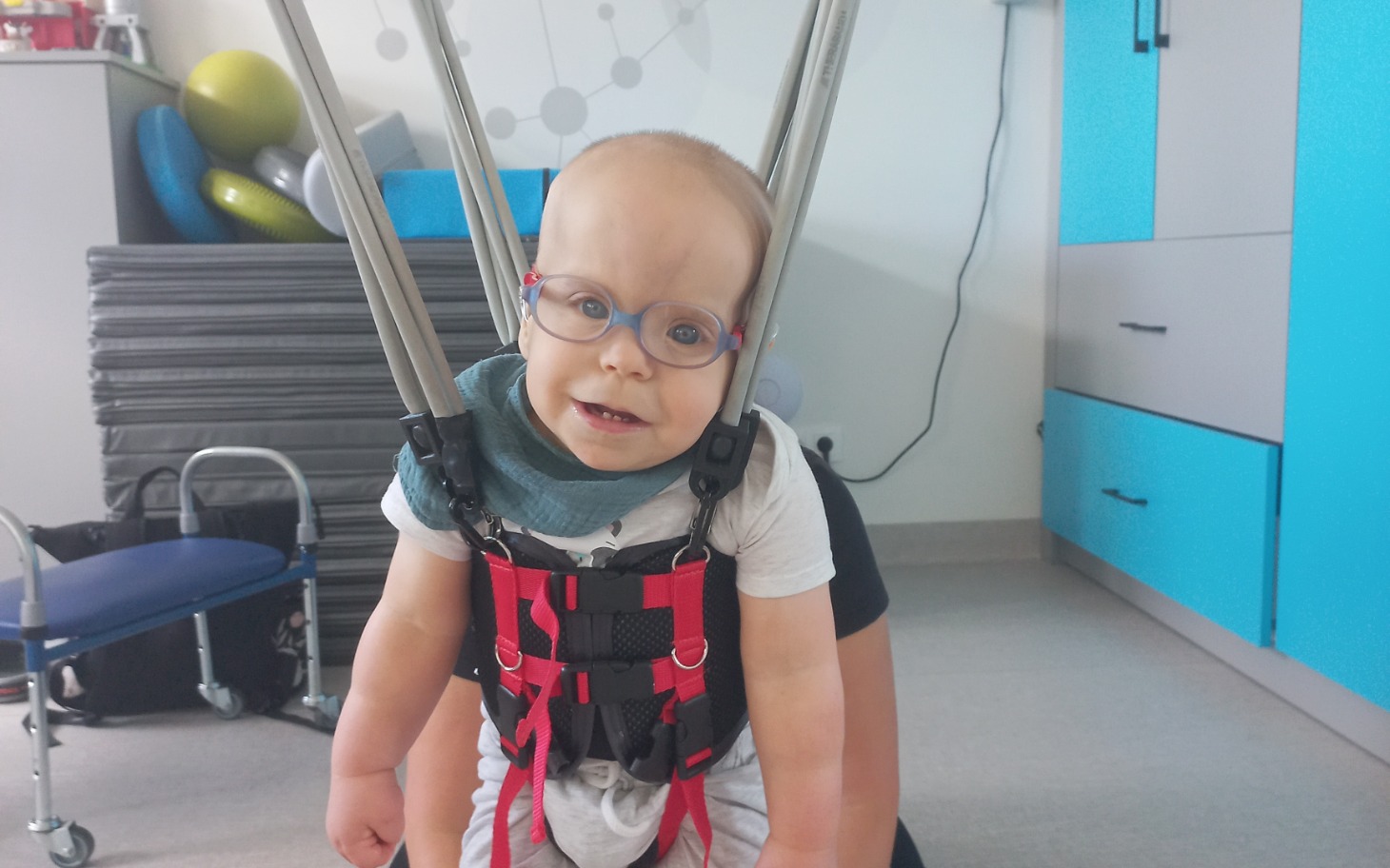 
        Małe dziecko w uprzęży terapeutycznej, uśmiechnięte i z okularami.
    