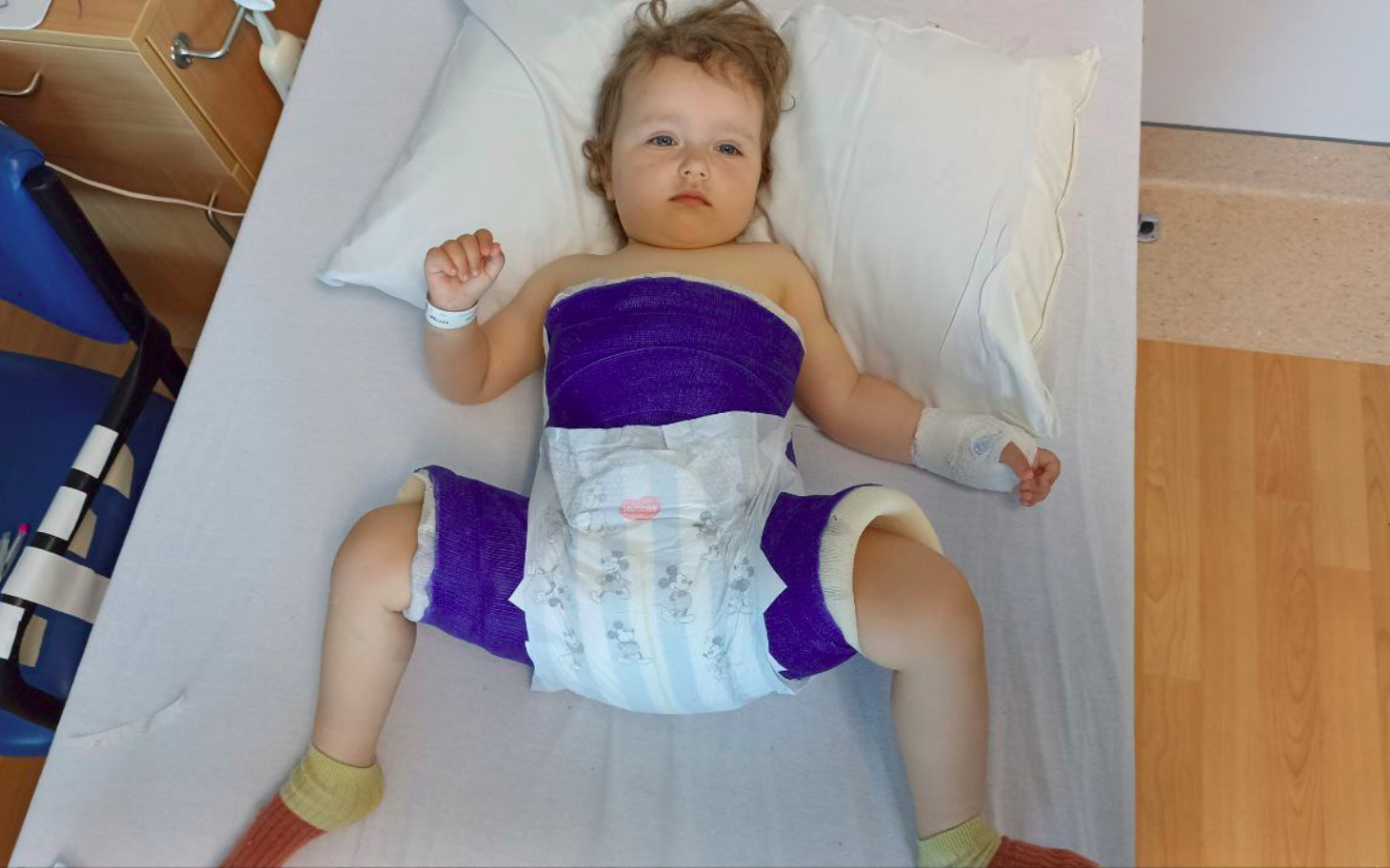 
        Dziecko w szpitalu, z opatrunkiem na ciele, leży na łóżku.
    