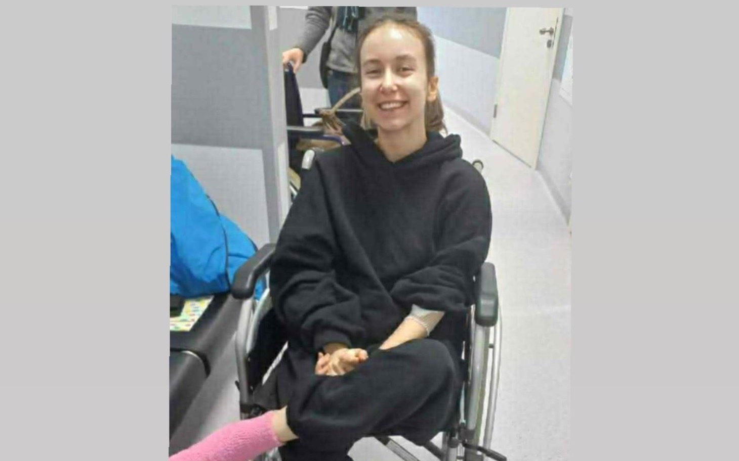 
        Młoda kobieta uśmiechająca się w wózku inwalidzkim, w szarym pomieszczeniu.
    