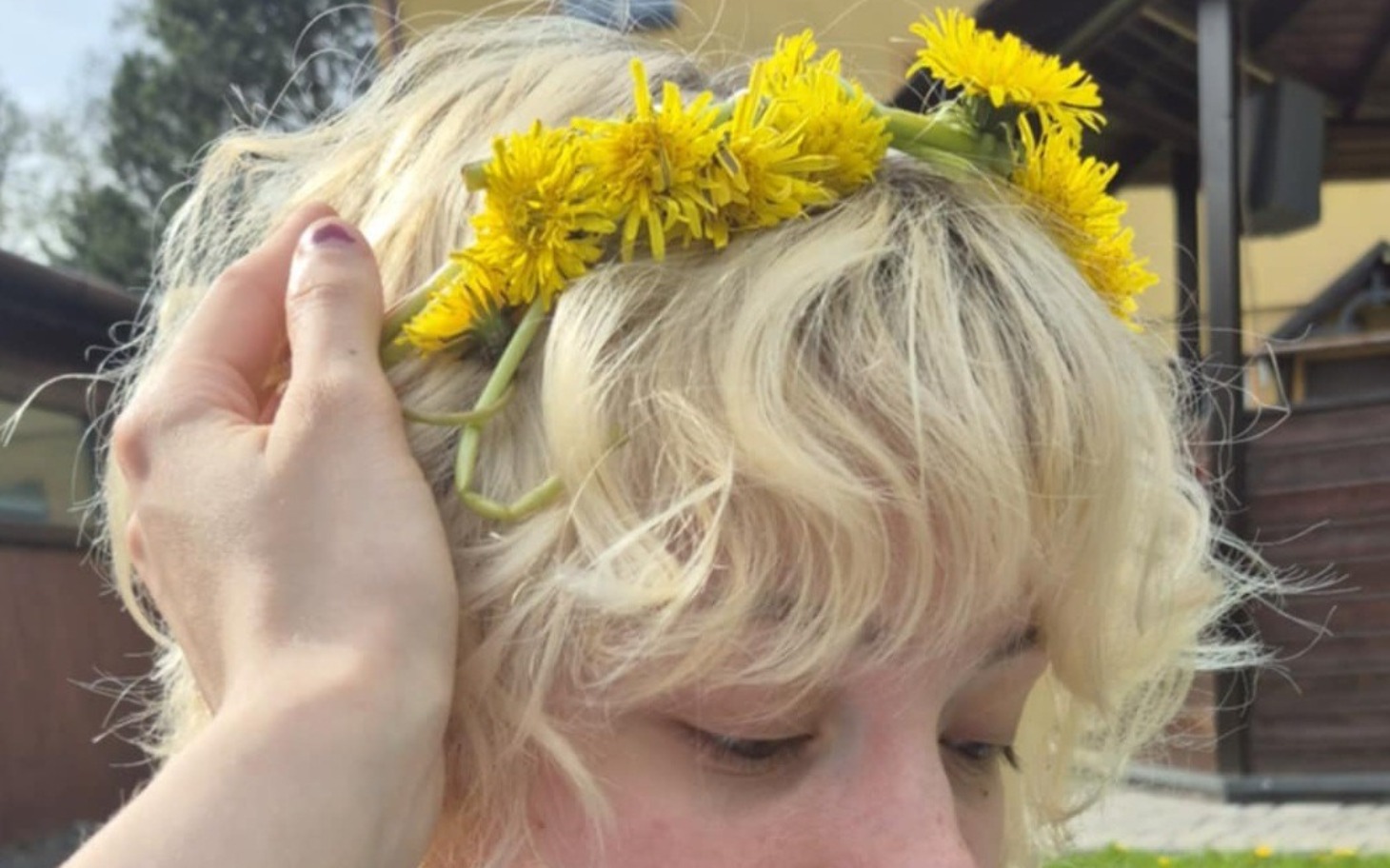 
        Kwiatowy wianek z mniszka na głowie, w tle wiosenna sceneria.
    