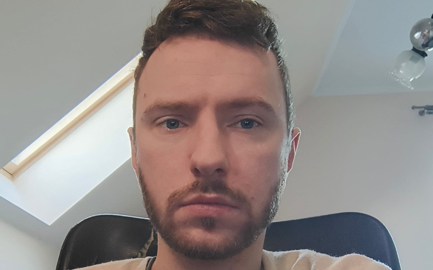 
        Mężczyzna z brodą siedzący przy biurku, z oknem w tle.
    