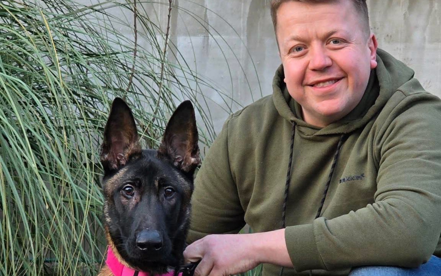 
        Mężczyzna z psem rasy owczarek niemiecki w otoczeniu zieleni.
    