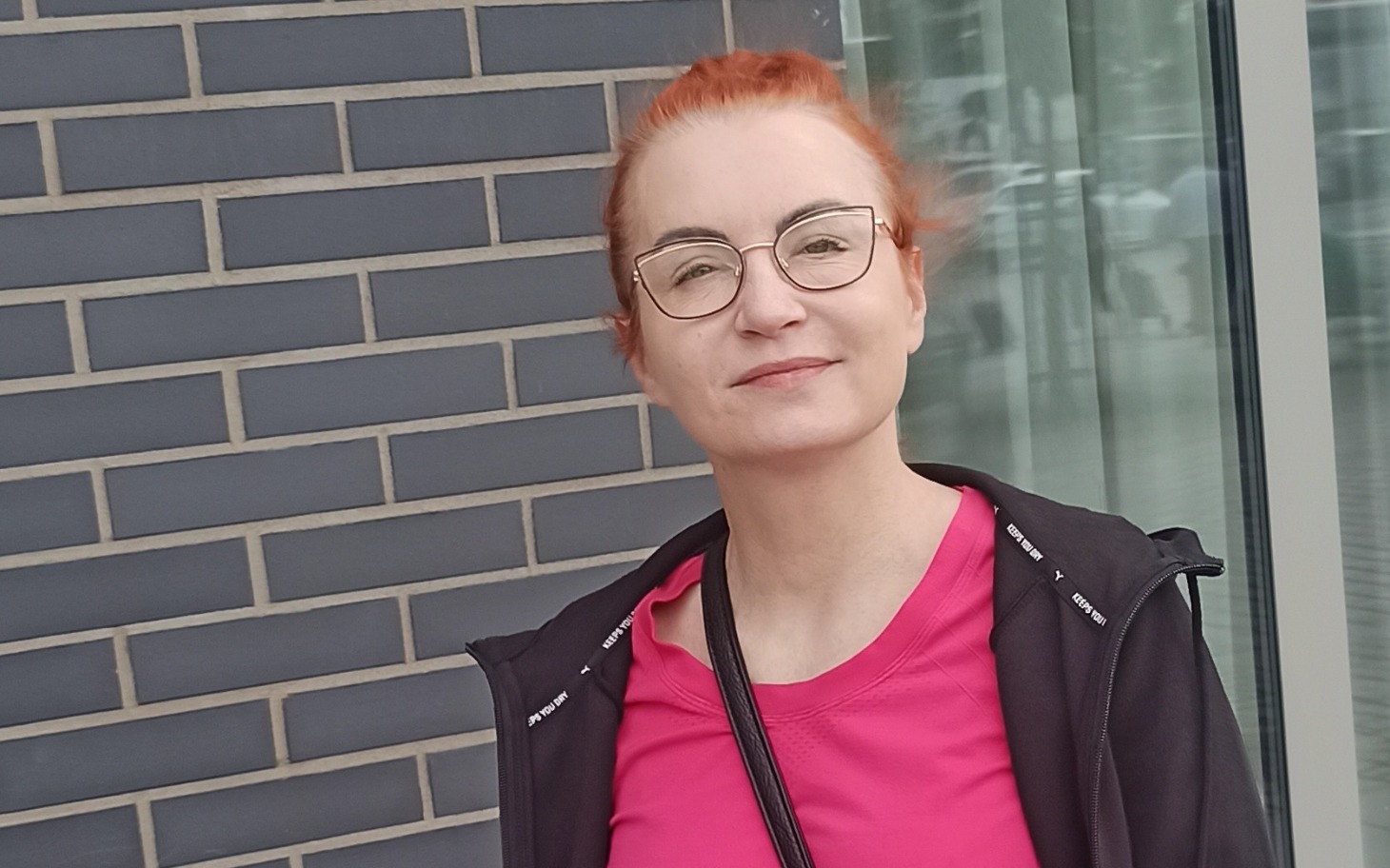 
        Kobieta w okularach, uśmiechająca się na tle ceglanej ściany.
    