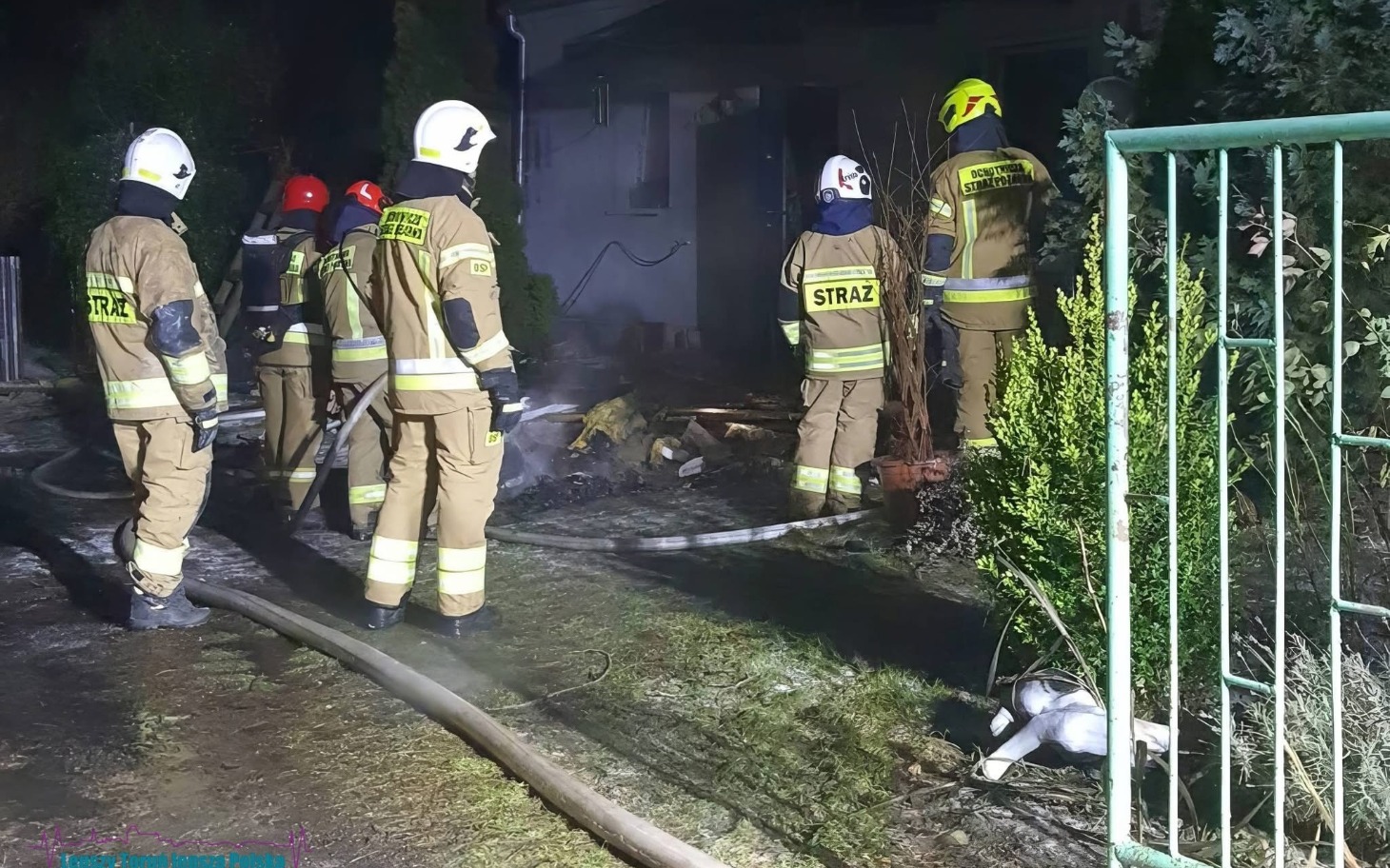 
        Strażacy gaszą pożar w nocy, otoczeni dymem i zniszczeniami.
    