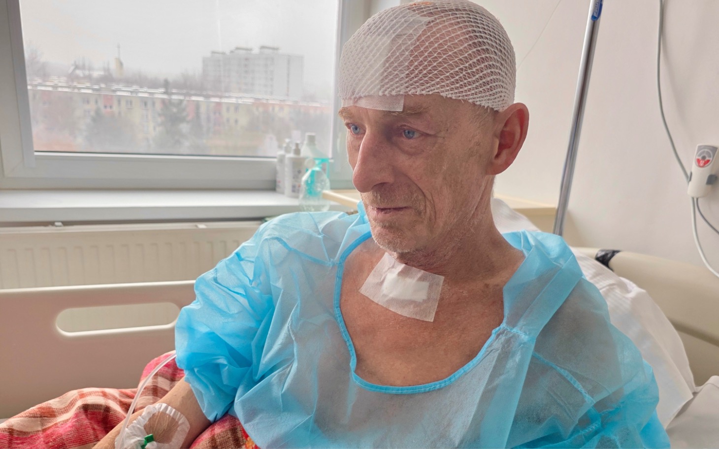 
        Mężczyzna w szpitalu, w niebieskim fartuchu, z opatrunkiem na szyi.
    