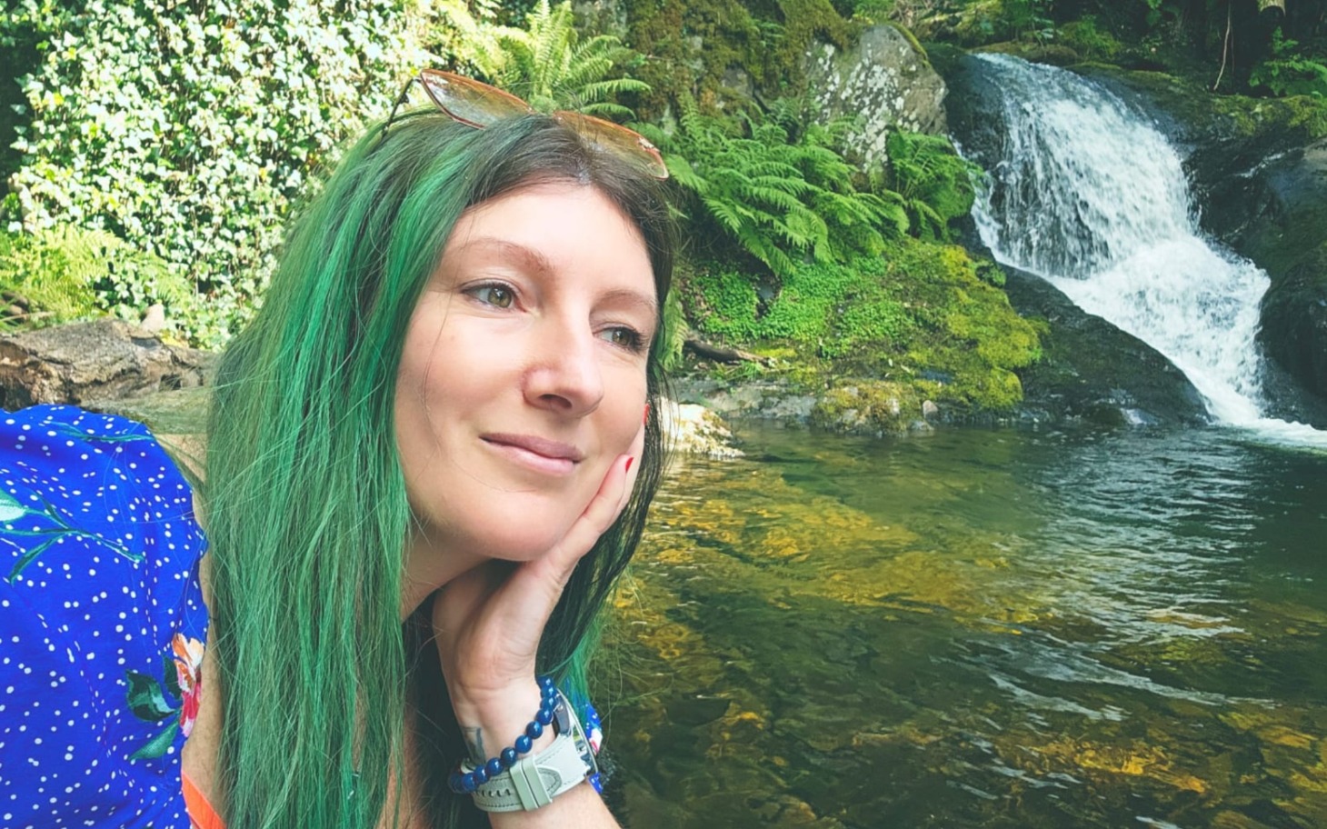 
        Kobieta z zielonymi włosami przy wodospadzie w lesie.
    