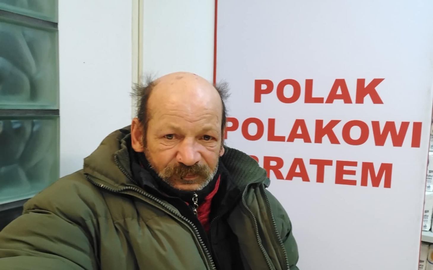 
        Mężczyzna w kurtce, pozujący przy plakacie.
    