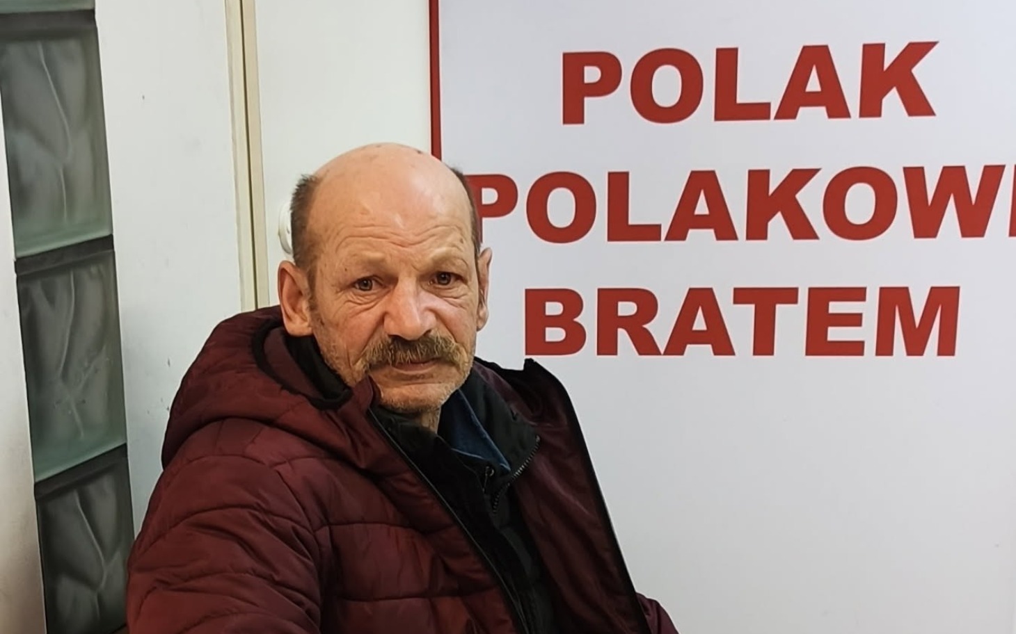 
        Mężczyzna w czerwonej kurtce siedzi przed napisem 'Polak Polakowi Bratem'.
    