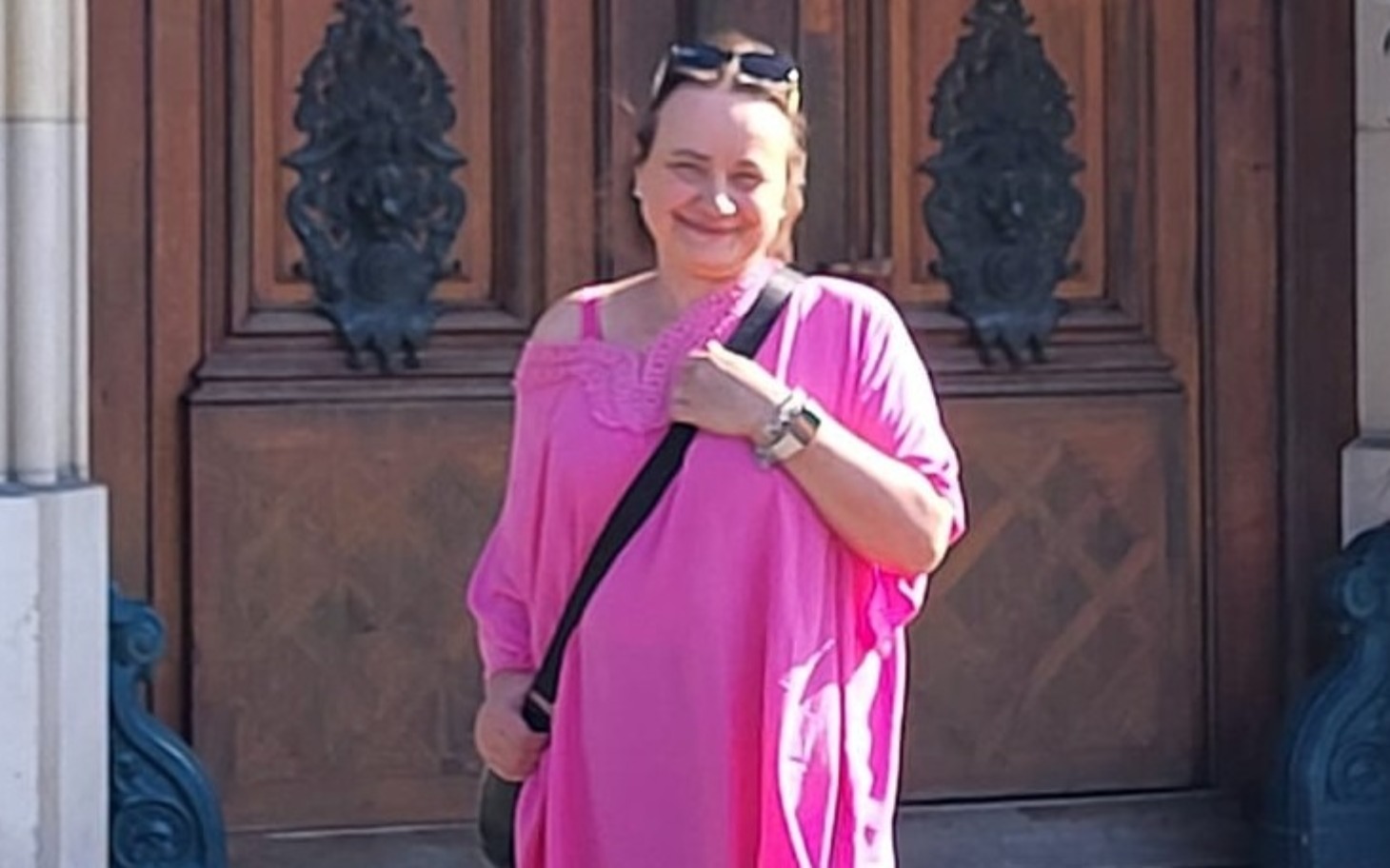 
        Kobieta w różowej sukience, uśmiechająca się przed drzwiami.
    