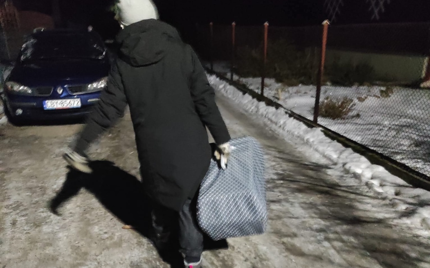 
        Osoba w czarnym płaszczu idąca nocą z torbą.
    