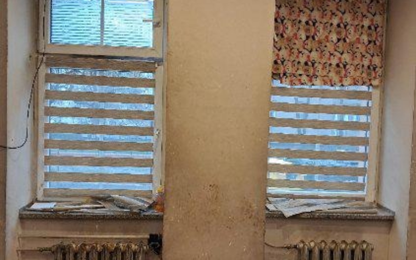 
        Dwa okna z roletami, z widocznymi zniszczeniami na ścianie.
    