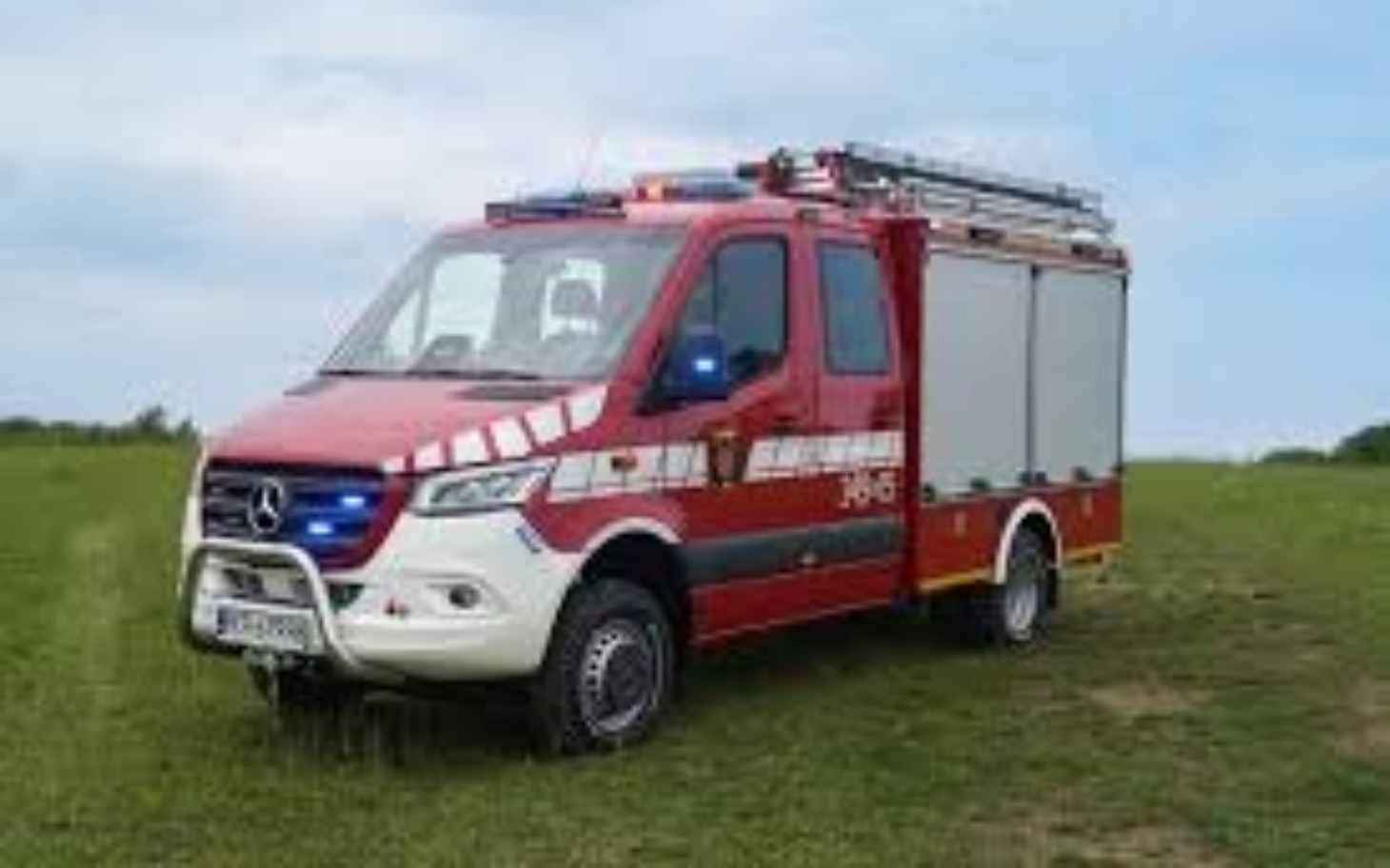 
        Wóz strażacki na tle zielonego terenu, gotowy do akcji.
    