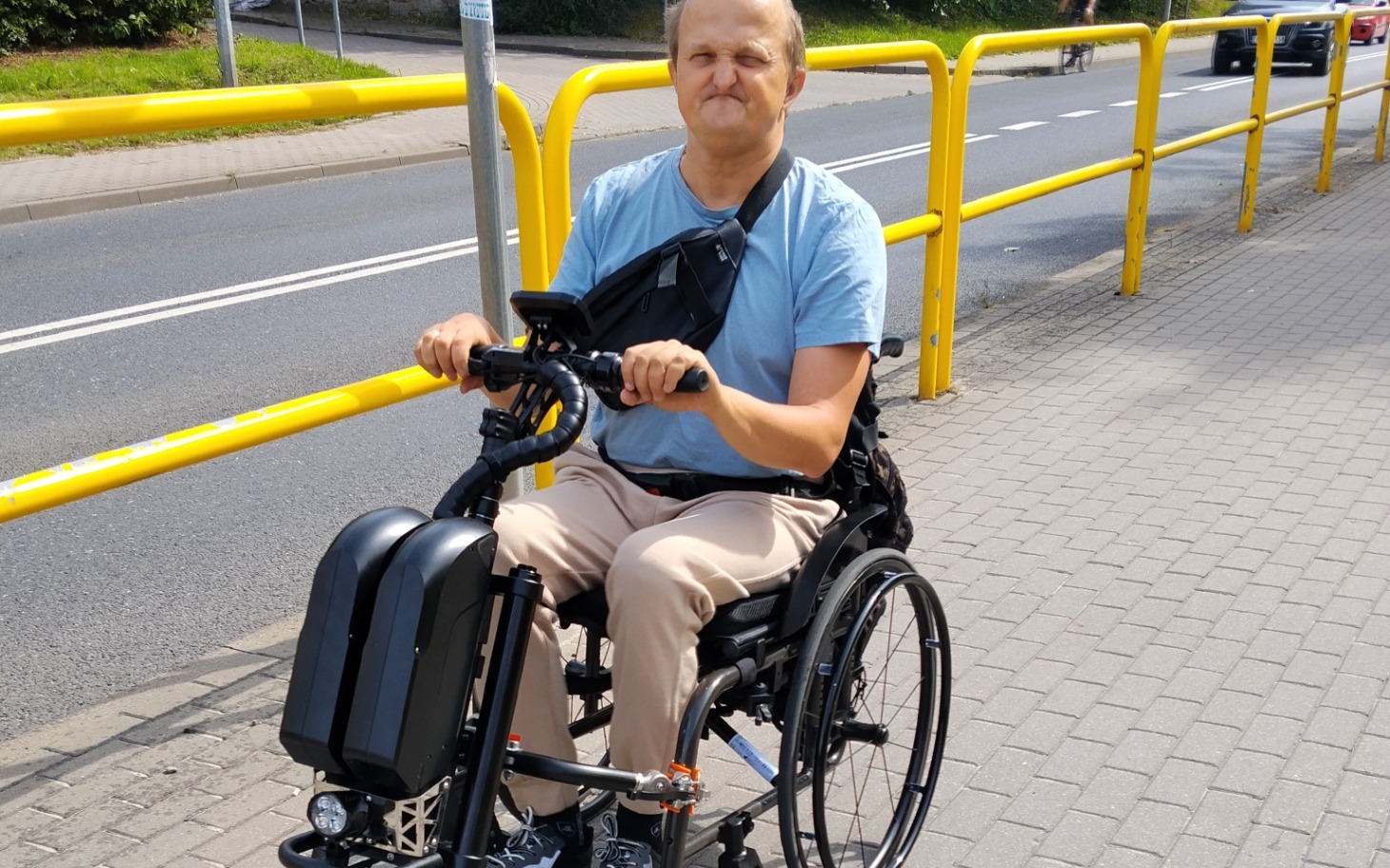 
        Osoba na wózku elektrycznym na ulicy z żółtym barierowym przęsłem, uśmiech i radosna atmosfera.
    