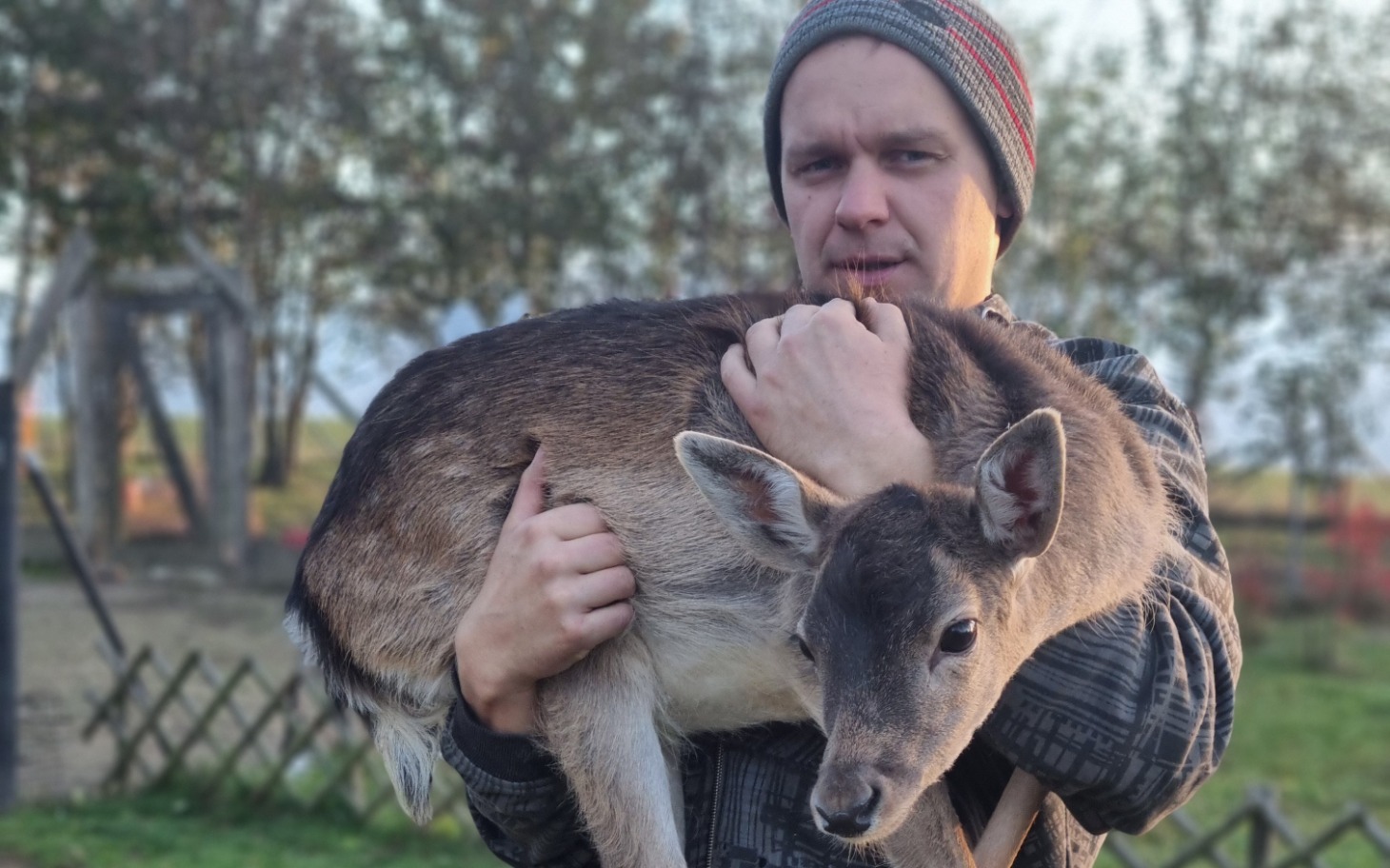 
        Mężczyzna trzymający sarnę w ramionach, na tle natury.
    