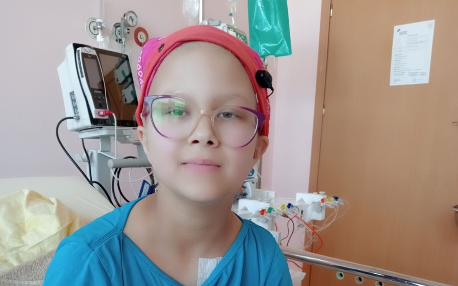 
        Dziewczynka w szpitalu z opaską na głowie i uśmiechem.
    