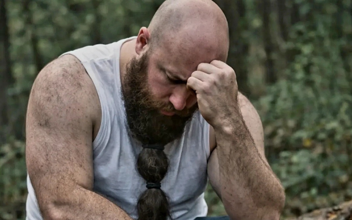 
        Mężczyzna z brodą siedzi w lesie, wygląda na zamyślonego.
    
