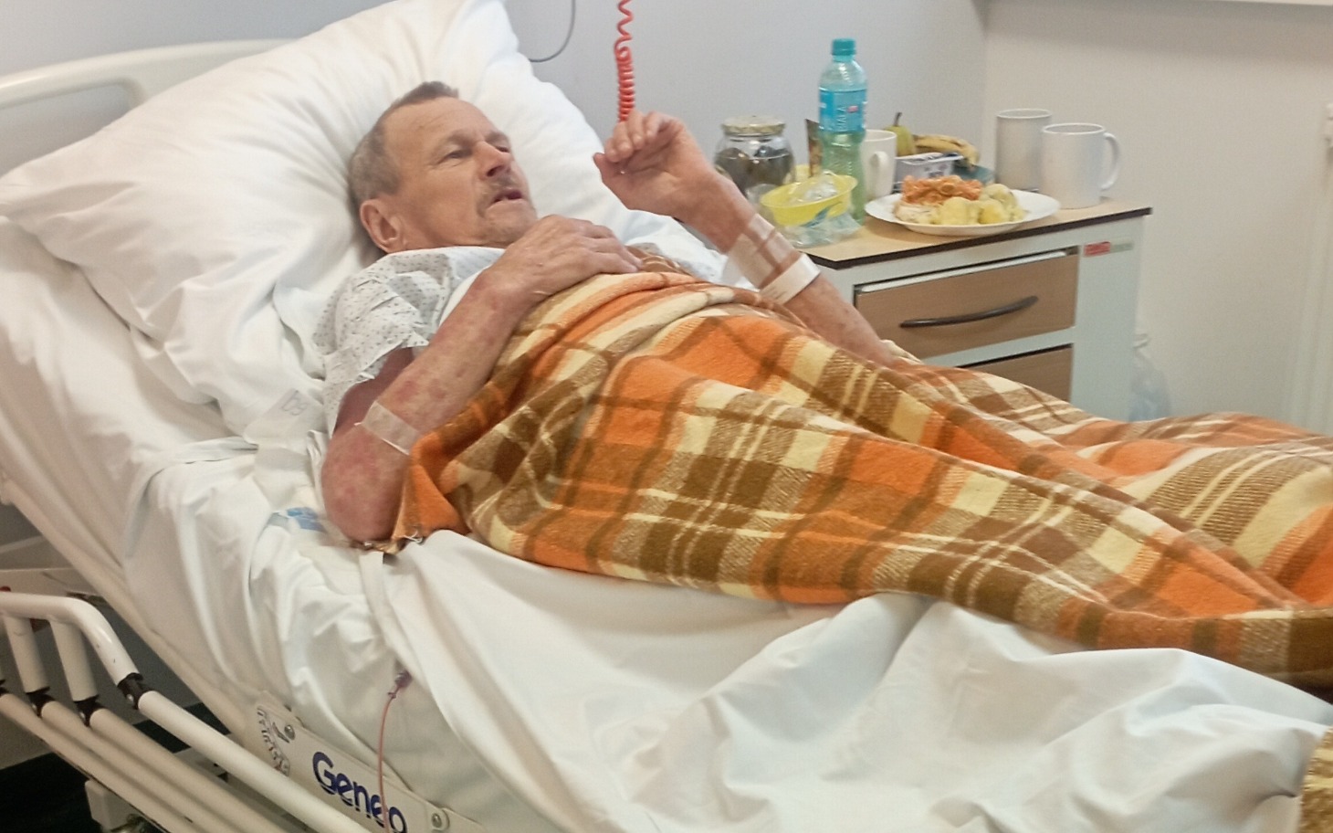 
        Starszy mężczyzna w szpitalnym łóżku, rozmawia z kimś.
    