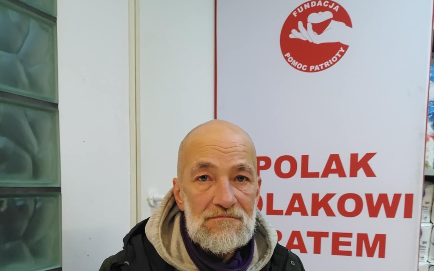 
        Mężczyzna z siwą brodą stoi przed bannerem fundacji.
    