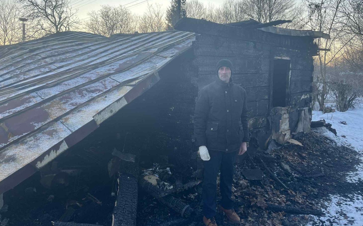 
        Mężczyzna stoi obok spalonego budynku w zimowej scenerii.
    