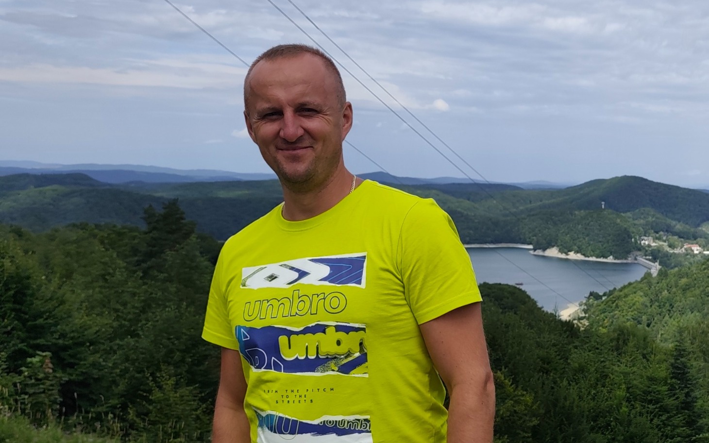 
        Mężczyzna w żółtej koszulce na tle górskiego krajobrazu.
    