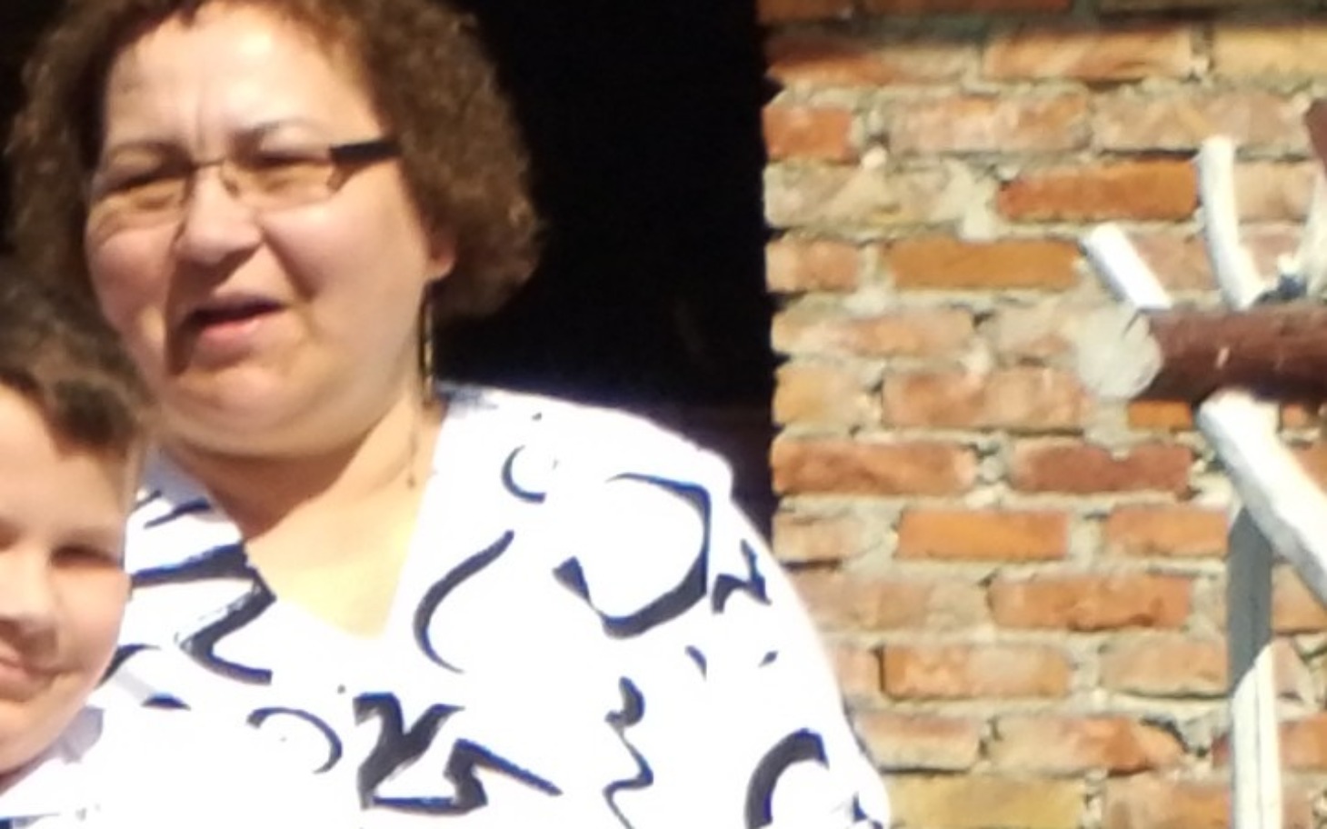 
        Kobieta z okularami, uśmiechająca się przy ceglanej ścianie.
    