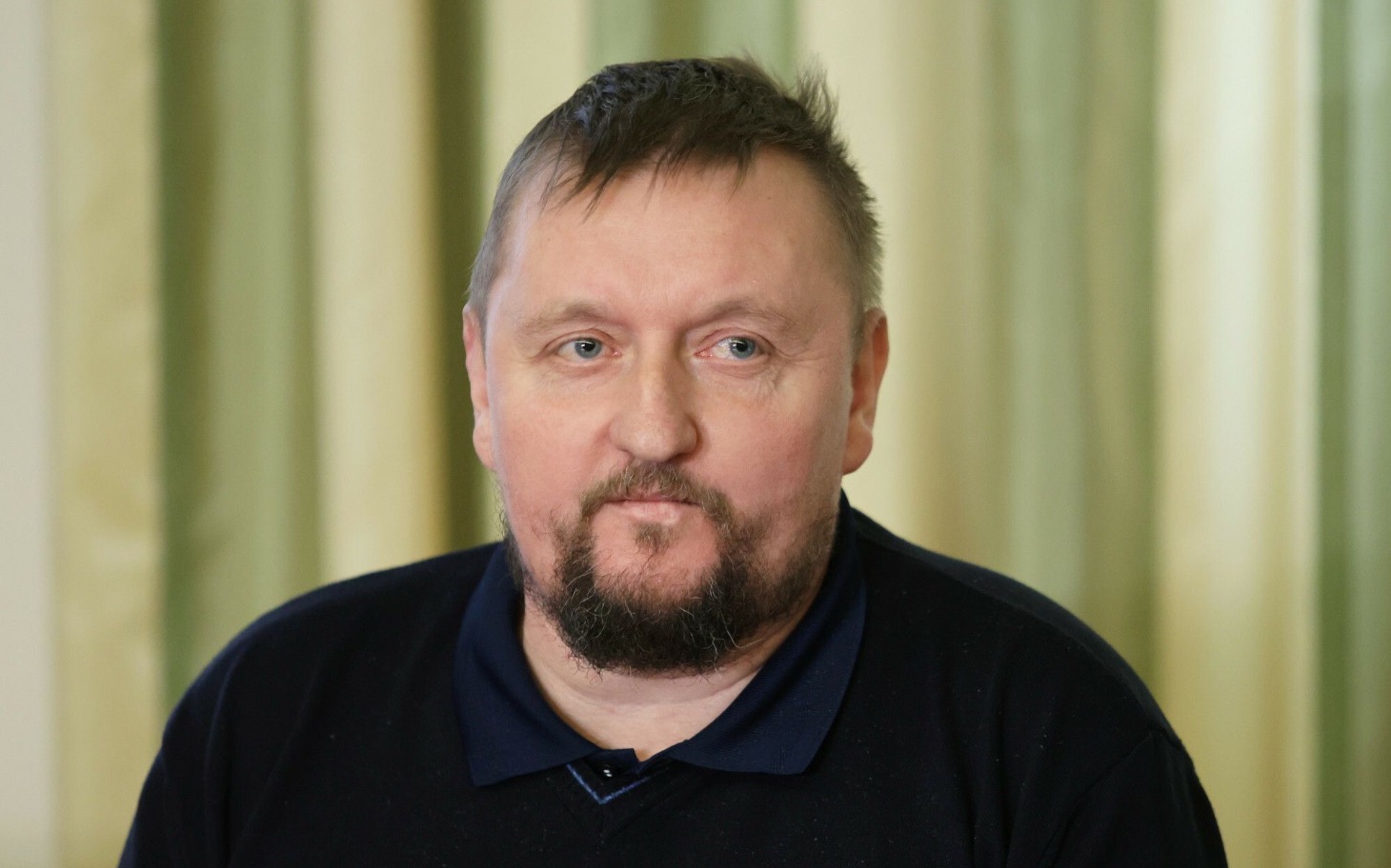 
        Mężczyzna z brodą w czarnym swetrze, siedzący w pomieszczeniu z zasłonami.
    