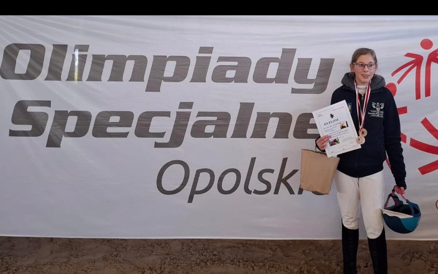 
        Młoda dziewczyna z medalami, trzymająca dyplom na tle banera Olimpiad Specjalnych.
    