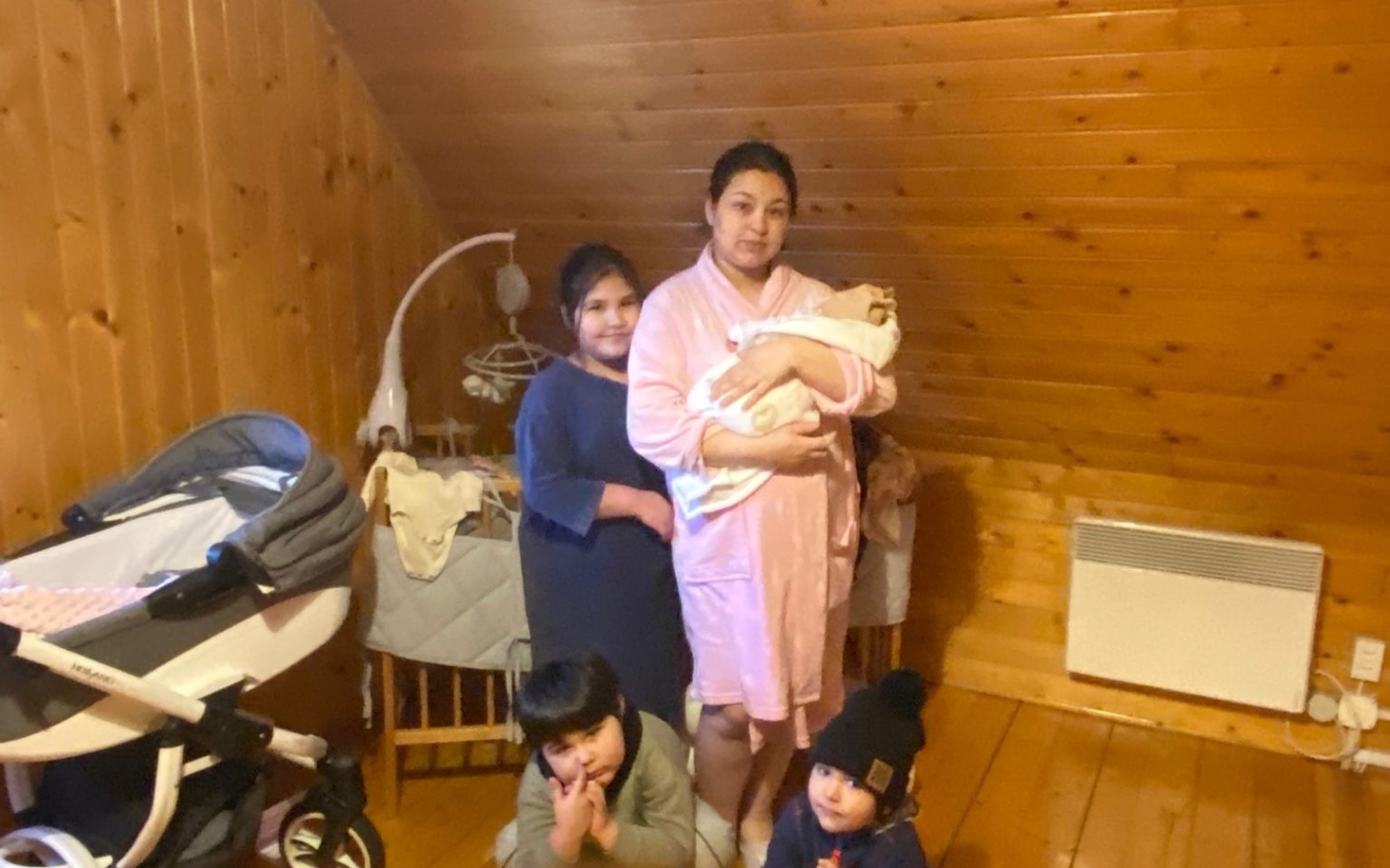 
        Rodzina z dziećmi w drewnianym wnętrzu, matka trzyma noworodka.
    