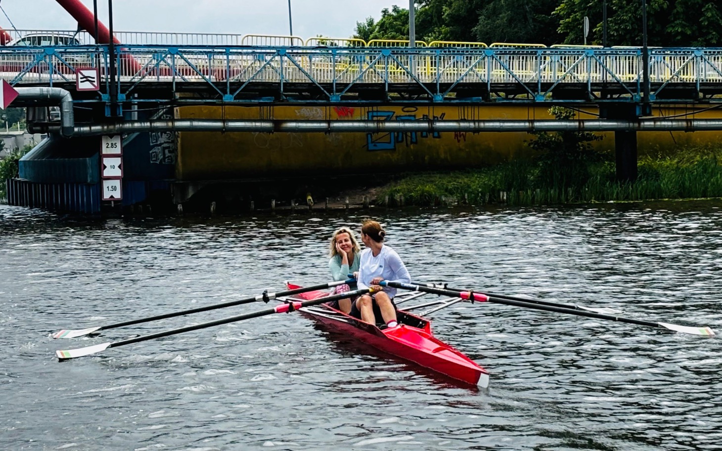 
        Dwie osoby w łódce wiosłowej na wodzie, w tle most.
    