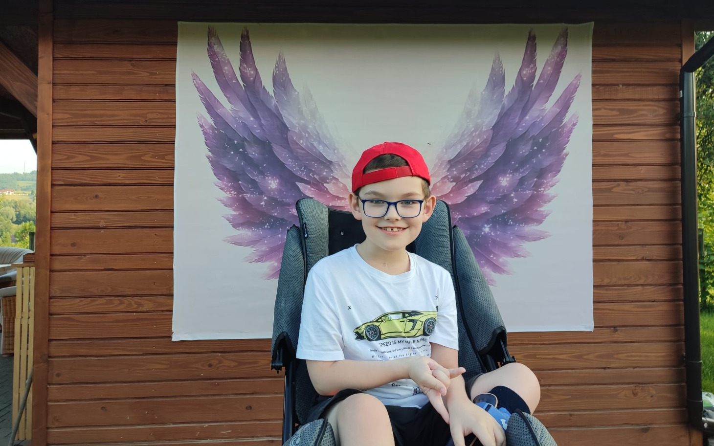 
        Chłopiec w czapce, siedzący na wózku przed tłem z anielskimi skrzydłami.
    