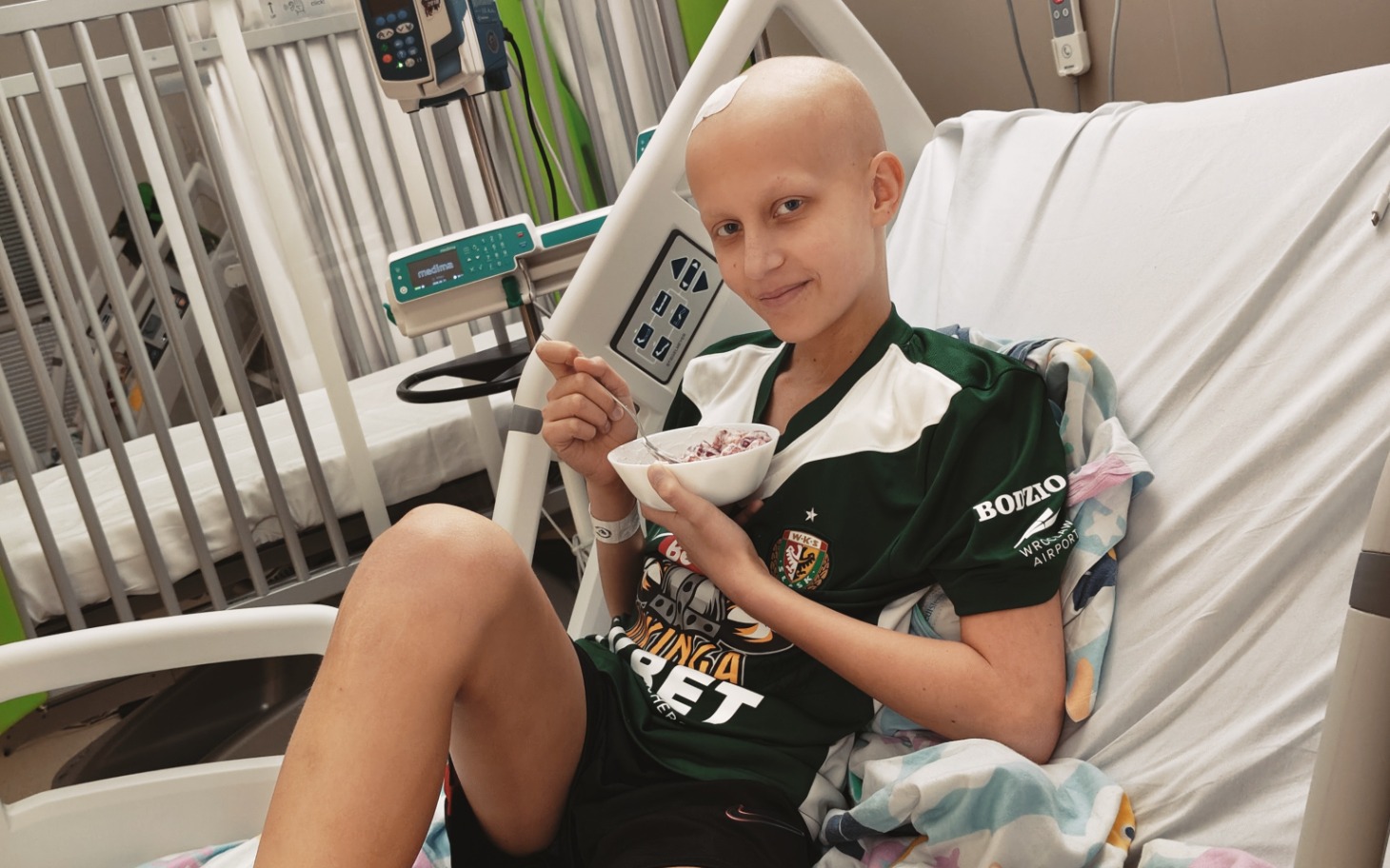 
        Młoda dziewczyna w szpitalu, jedząca deser w koszulce sportowej.
    