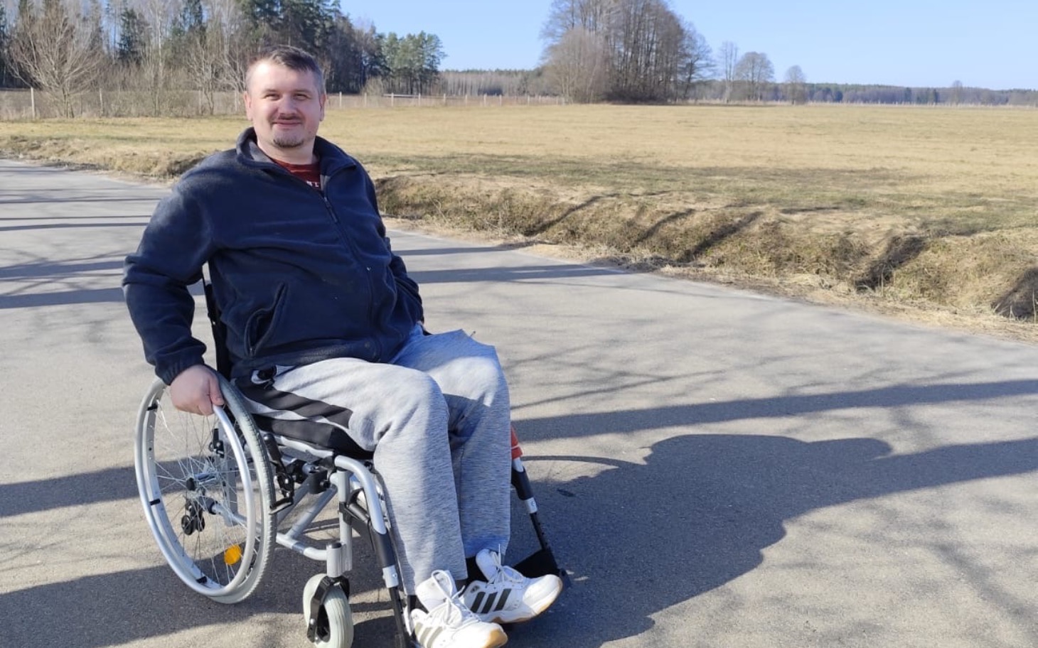 
        Mężczyzna na wózku inwalidzkim, uśmiechający się na tle natury.
    