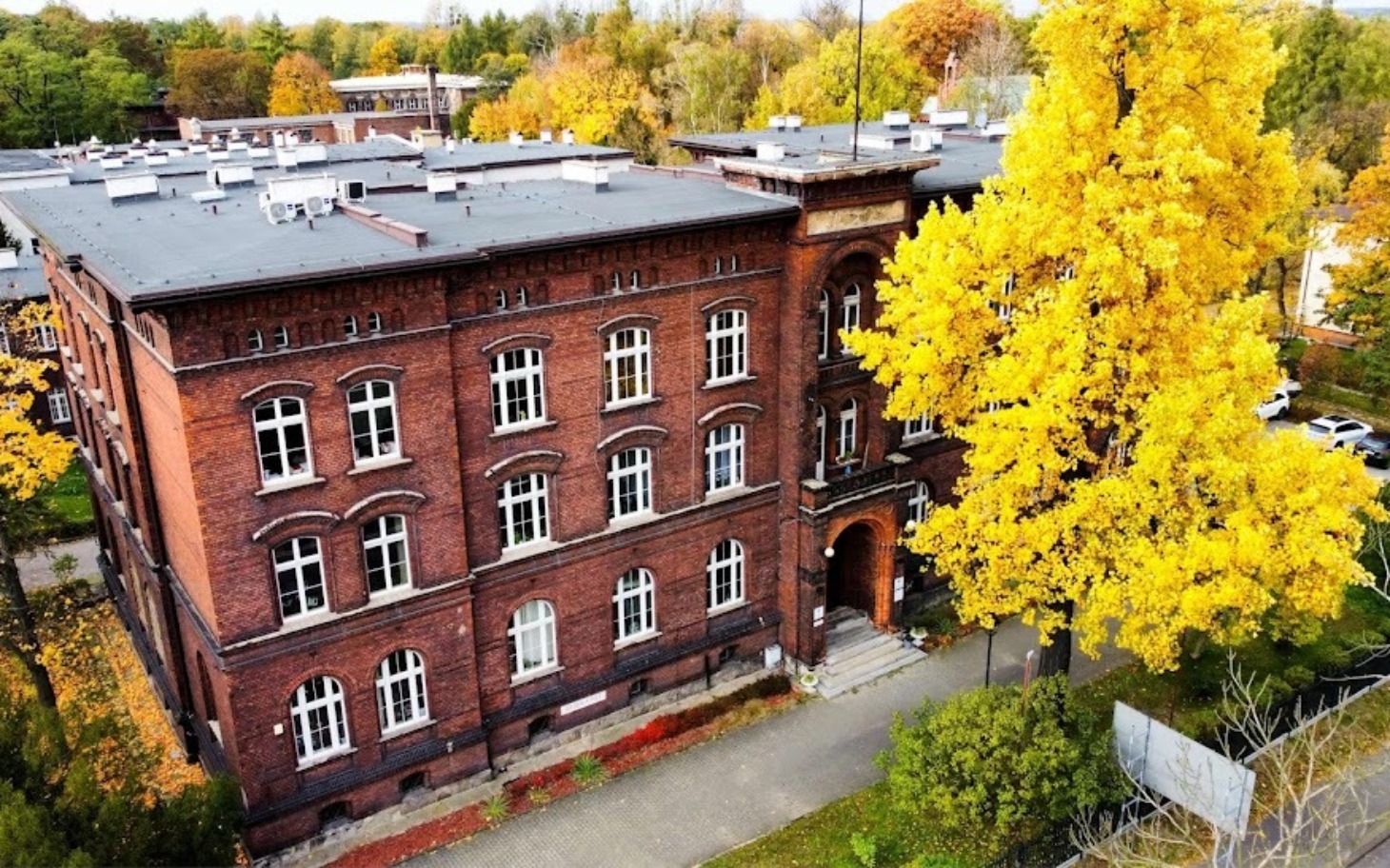
        Widok na stary budynek otoczony drzewami w jesiennych kolorach.
    