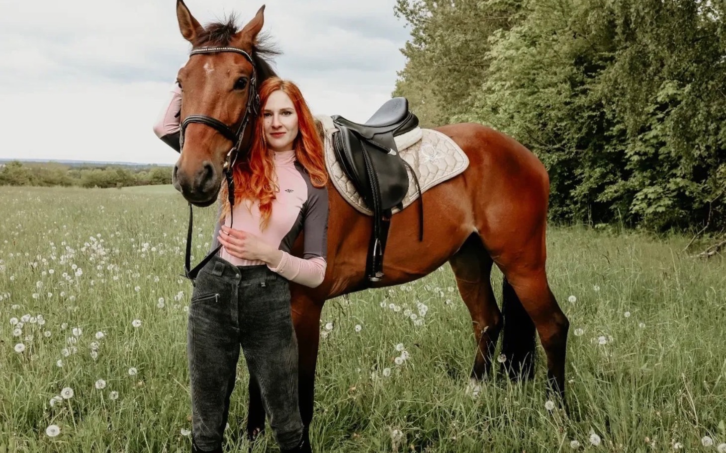 
        Kobieta z rudymi włosami pozująca z koniem na łące.
    