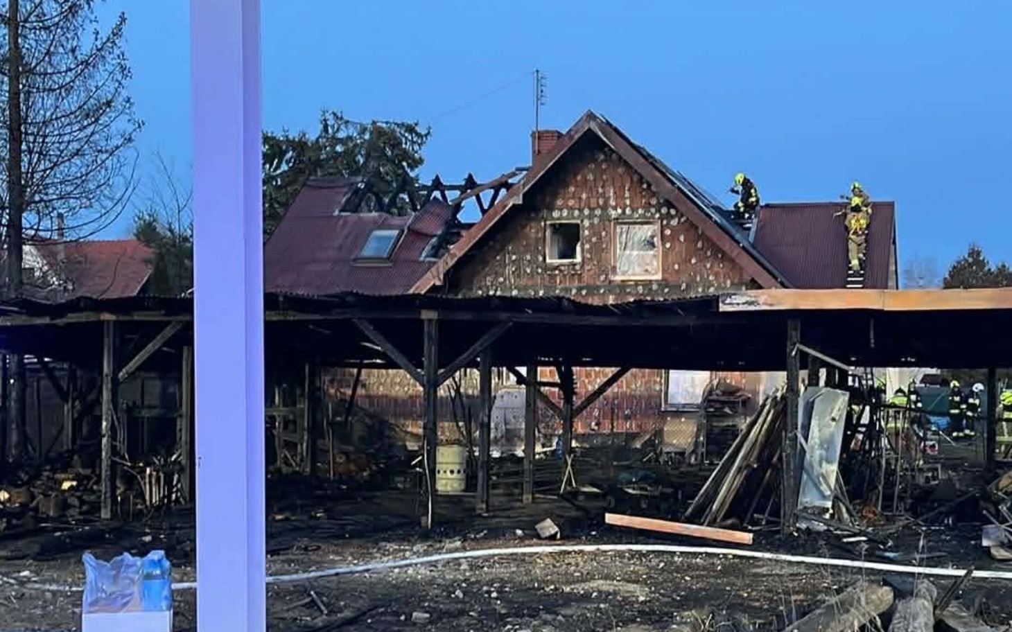 
        Zniszczony budynek po pożarze, strażacy na dachu.
    