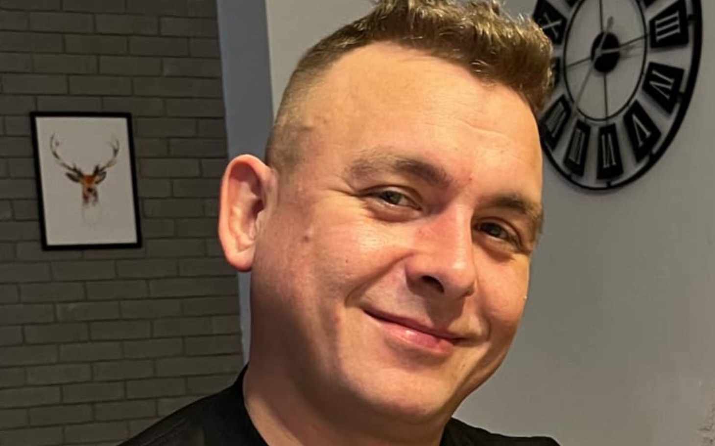 
        Mężczyzna uśmiechający się w domu, z zegarem na ścianie.
    