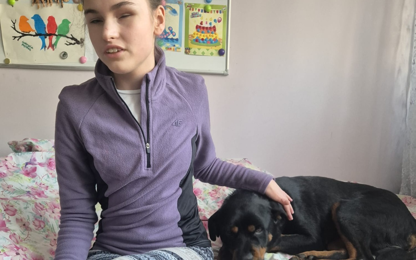 
        Dziewczyna siedząca na łóżku z psem obok, w fioletowej bluzie.
    