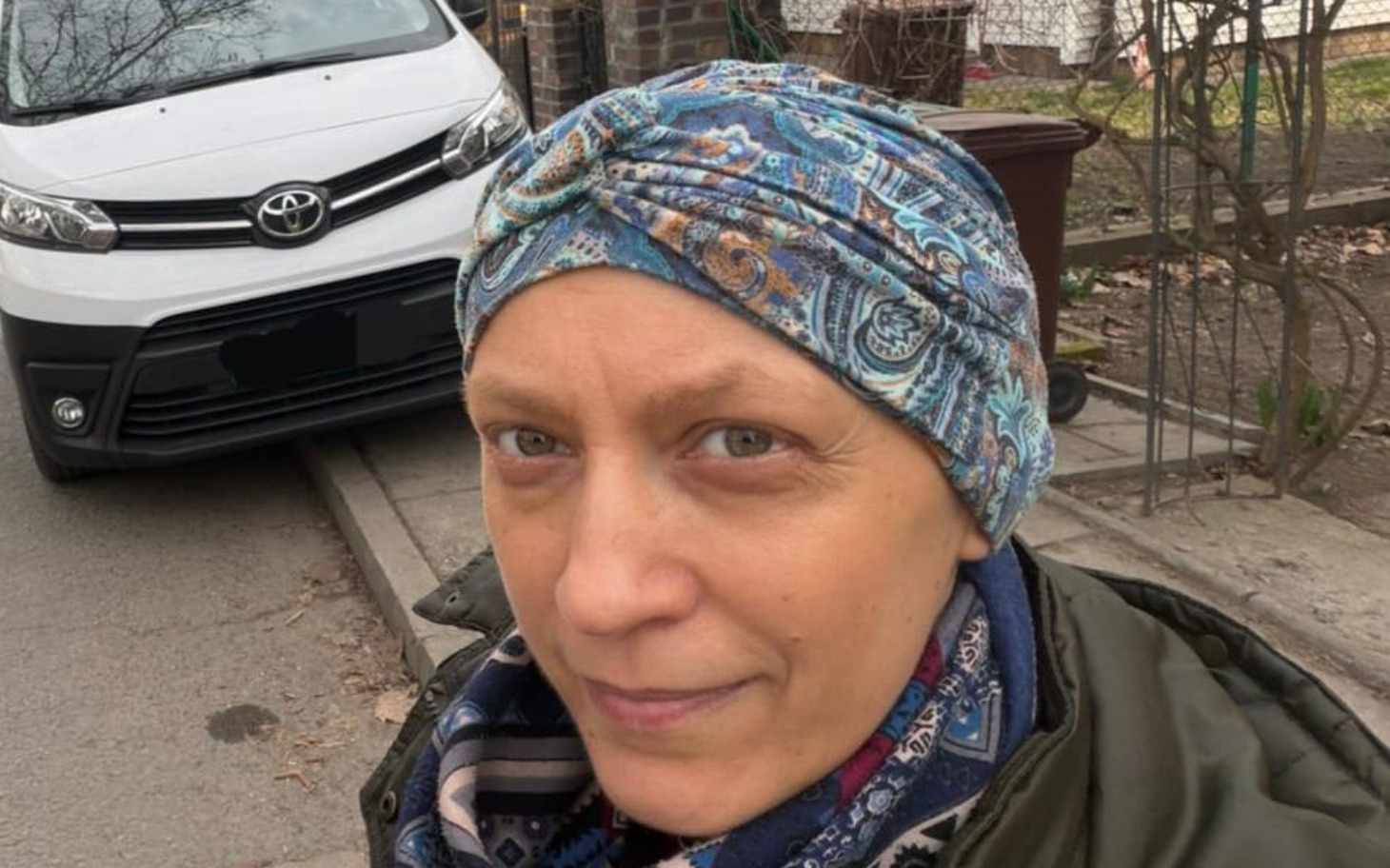 
        Kobieta w chustce, uśmiechająca się do kamery, na tle samochodu.
    