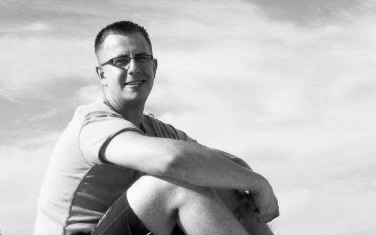 
        Mężczyzna siedzący na plaży, uśmiechający się do kamery.
    