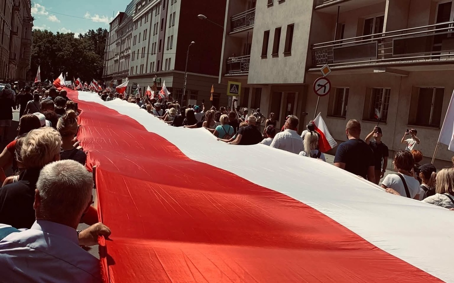 
        Manifestacja z flagą Polski, tłum ludzi na ulicy.
    