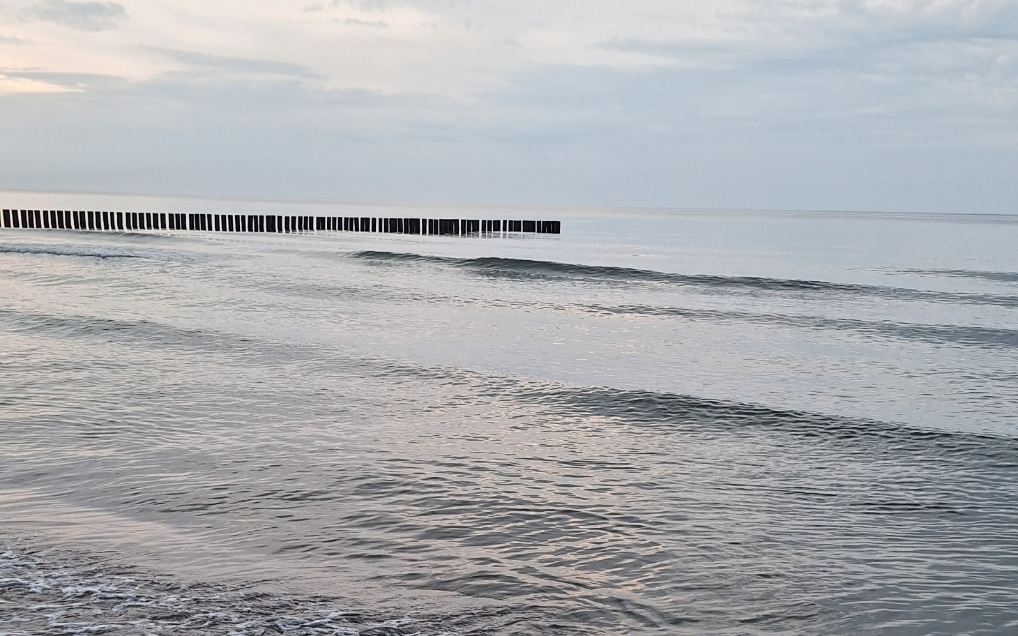 
        Spokojna plaża z falami, drewniane pomosty w oddali.
    