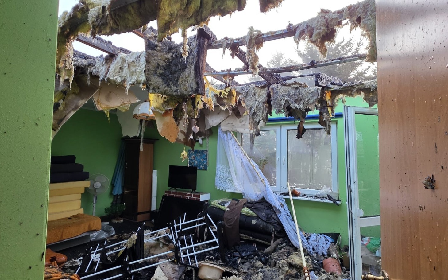 
        Zniszczone wnętrze domu po pożarze, z uszkodzonym dachem i meblami.
    