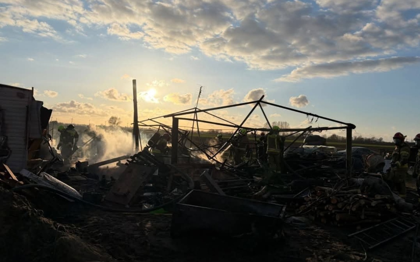 
        Scena pożaru, strażacy pracują w zgliszczach pod wieczornym niebem.
    